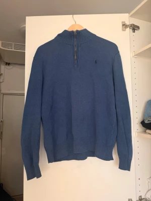 Blå stickad tröja Ralph Lauren - Snygg blå stickad tröja från Polo Ralph Lauren med halv dragkedja och hög krage. Tröjan har ribbade muddar vid ärmslut och nederkant samt den klassiska lilla loggan broderad på bröstet. Perfekt till jeans eller chinos.