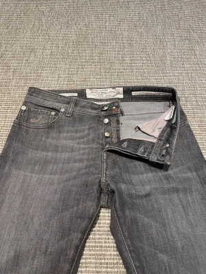 Gråa jeans från Jacob Cohen - Snygga grå jeans från Jacob Cohen med klassisk femficksmodell och knappgylf. Jeansen har diskreta slitningar, kontrastsömmar och en brun patch med logga bak i midjan. Perfekta för en avslappnad men stilren look.