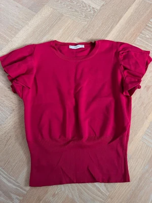 Röd topp med volangärmar från Mango - Snygg mörkrosa topp från Mango med volangdetaljer på ärmarna och rund halsringning. Modellen är ribbad nedtill för en smickrande passform och har korta ärmar med extra volym. Perfekt för dig som vill ha en färgstark och trendig look. Storleken är mer som S
