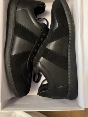 Maison Margiela Gats - Stilrena svarta sneakers med ovandel i både läder och mocka. Skorna har klassisk låg profil, svarta skosnören och en slitstark gummisula med mönster undertill. Perfekta för dig som gillar minimalistisk och clean stil.