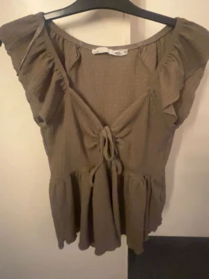Beige volangtopp med knyt - Supersöt beige topp med volangdetaljer och knyt framtill. Toppen har en luftig passform och är tillverkad i ett mjukt, lätt material som känns skönt mot huden. Perfekt för dig som gillar en avslappnad men ändå trendig stil.