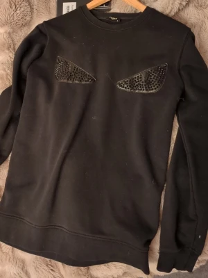 Svart sweatshirt från Fendi med nitar - (OBS 2 bitar saknas på varje öga) Cool svart sweatshirt från Fendi med två ögonformade detaljer på bröstet, dekorerade med svarta nitar för en edgy look. Tröjan har rund halsringning och är tillverkad i mjukt bomullsmaterial. Perfekt för dig som vill sticka ut med en unik design.