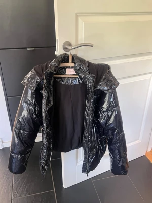 Svart glansig pufferjacka med huva - Säljer en svart, glansig pufferjacka med huva och dragkedja framtill. Jackan har en oversized look och breda kanaler som ger en riktigt puffig känsla. Perfekt för kalla dagar när du vill hålla dig varm.