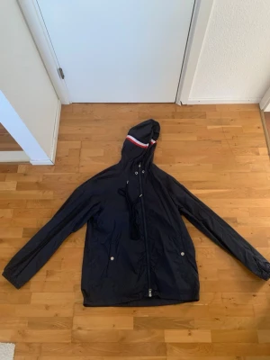 Svart vindjacka med huva från Moncler - Snygg svart vindjacka från Moncler med huva och dragkedja framtill. Jackan har vita och röda detaljer på huvan, två fickor med tryckknappar och justerbar dragsko i midjan. Tillverkad i lätt, vattenavvisande material som passar perfekt för blåsiga dagar.