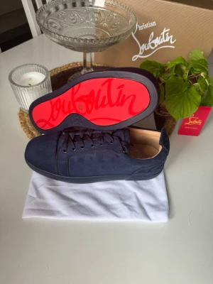 Mörkblå sneakers från Christian Louboutin - Snygga mörkblå sneakers från Christian Louboutin i mocka med klassisk röd sula och diskret logga på sidan. Skorna har lågt skaft, matchande mörkblå snören och en ikonisk design som sticker ut med den röda undersidan.