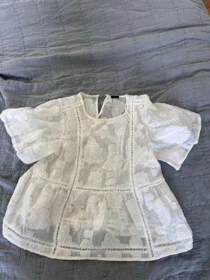 Vit blommig blus med puffärm - Söt vit blus med blommigt mönster och puffiga korta ärmar. Blusen har transparenta partier och dekorativa sömmar som ger en romantisk känsla. Rund halsringning med knapp i nacken. Perfekt för en trendig och luftig look.