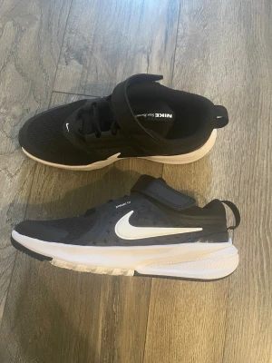 Svarta Nike sneakers med vit sula - Snygga svarta Nike sneakers med vit sula och vit swoosh på sidan. Skorna har meshmaterial för bra ventilation, kardborreband och elastiska snören för enkel påtagning. Perfekta för en sportig och avslappnad stil.