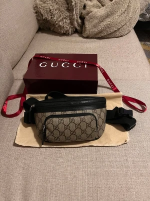 GG Supreme bälte väska - Säljer en snygg  GG Supreme bälte väska i beige och brunt med klassiskt GG-mönster. Väskan har svart justerbart band, dragkedja och en mindre ficka framtill. Perfekt accessoar för dig som vill ha både stil och funktion.