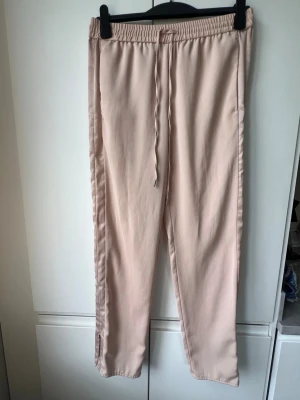 Beigerosa loungewear byxa med revärer - Säljer ett par ljusbeige byxor med resår och dragsko i midjan. Byxorna har raka ben och dragkedjor längst ner vid bensluten för en snygg detalj. Glansigt tyg i revärer på benens sidor som snygg detalj. Loungewear. 