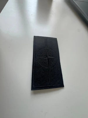 Stone island badge - black - Säljer en svart tygpatch från Stone Island med det klassiska kompasslogotypen broderad ton-i-ton. Rektangulär form och stilren design, perfekt att sy eller fästa på jackor, väskor eller kepsar för en unik look.