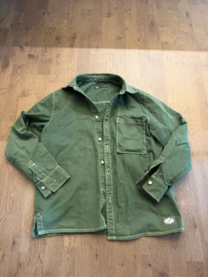 Grön overshirt från Zara - Stilren grön overshirt från Zara i kraftigt bomullstyg. Skjortan har klassisk krage, bröstficka och vita knappar framtill samt vid ärmsluten. Perfekt lager-på-lager-plagg med avslappnad passform och diskret patch nertill.