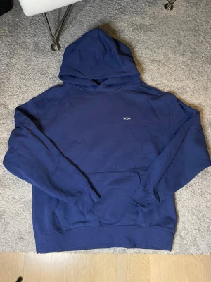 Mörkblå hoodie från MODA - En stilren mörkblå hoodie från MODA med klassisk huva och magficka. Tröjan har en enkel design med liten vit logga på bröstet och ribbade muddar vid ärmar och nederkant. Perfekt för en avslappnad look och tillverkad i mjukt bomullsmaterial.