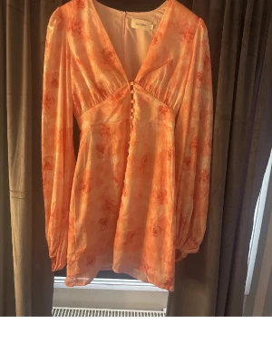 Blommig kortklänning från Adoore - Supersöt kortklänning från Adoore i ljus orange och rosa toner med blommigt mönster. Klänningen har v-ringning, långa puffärmar och dekorativa knappar framtill. Tillverkad i ett lätt, glansigt material som känns lyxigt och somrigt. Använd en gång. En knapp har gått av, men går lätt att sy fast igen! Har även klippt upp klänningen men den ser exakt ut som den korta. Pris går att diskutera.