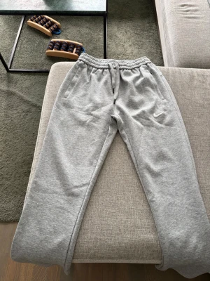 Gråa mjukisbyxor från Nike - Snygga gråa mjukisbyxor från Nike med broderad vit logga på benet. Byxorna har resår i midjan med dragsko och två sidofickor. Tillverkade i mjukt och bekvämt material som passar perfekt för chill eller träning.