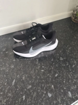 Svarta Nike sneakers med vit sula - Snygga svarta Nike sneakers med vit sula och grå swoosh på sidan. Ovandelen är i meshmaterial för bra andningsförmåga och komfort. Skorna har klassisk snörning och sportig design, perfekta för en aktiv stil. Reflekterande detaljer och lättviktssula för extra komfort.