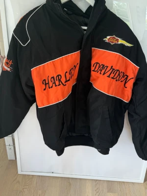 Harley Davidson svart och orange vindjacka - Cool vindjacka från Harley Davidson i svart och orange med stora broderade loggor och text på bröstet och ärmen. Jackan har dragkedja framtill, ståkrage och muddar vid ärmslut och midja. Perfekt för dig som gillar streetwear och motorcykelstil.