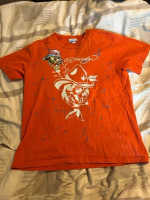 Orange Ed Hardy t-shirt med tryck - Cool orange t-shirt från Ed Hardy med stort vitt tryck av en dödskalle med cowboyhatt på framsidan. T-shirten har även ett färgglatt tryck av en bulldog och blå färgstänk. Klassisk passform och rund halsringning. Trycket är lite slitet men är fortfarande fin.