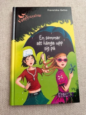 Vampyrsystrar: En sommar att hänga upp sig på - Rolig och spännande bok i serien Vampyrsystrar av Franziska Gehm. Omslaget är färgstarkt med illustration av två tjejer under ett svart paraply mot en gulgrön bakgrund. Inbunden bok, perfekt för dig som gillar äventyr och humor med övernaturliga inslag.