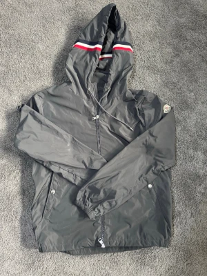 Moncler grimpeur  - Snygg grå vindjacka från Moncler med huva och dragkedja. Jackan har vita och röda ränder på huvan samt Moncler-logga på ärmen. Två fickor med tryckknappar och justerbar dragsko i huvan. Tillverkad i lätt och tåligt material, perfekt för blåsiga dagar.