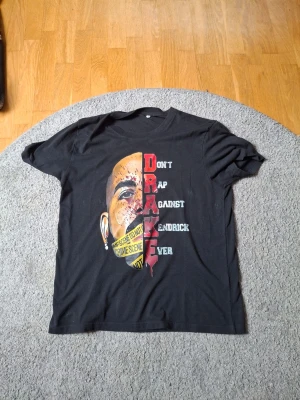 Svart t-shirt med Drake-tryck - Cool svart t-shirt med stort tryck av Drake på framsidan och texten 'Don't Rap Against Kendrick Ever'. Motiv med ansikte och gul tejp med 'Crime Scene'. Klassisk passform och rund hals. Perfekt för dig som gillar hiphop och streetwear.