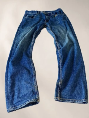 Blå baggy jeans med raka ben - Säljer ett par klassiska blå jeans med baggy passform och raka ben. Jeansen har fem fickor, normal midja och slitningar för en cool vintage-look. Tillverkade i kraftig denim, perfekta för en avslappnad stil.