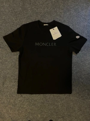Svart Moncler t-shirt med logga - Snygg svart t-shirt från Moncler med klassisk logga tryckt på bröstet och en patch på ärmen. T-shirten har rund hals och korta ärmar, tillverkad i mjuk bomull för en skön känsla. Perfekt för dig som gillar stilrena och exklusiva plagg.Finns även i navy blå och Beige. Storlek L