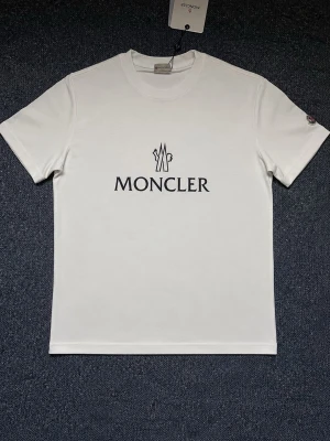 Vit Moncler t-shirt med logga - Snygg vit t-shirt från Moncler med klassisk svart logga tryckt på bröstet. T-shirten har rund hals, korta ärmar och en stilren passform. Tillverkad i mjuk bomull för maximal komfort. Perfekt för dig som gillar exklusiva och enkla plagg. Skriv för fler bilder. Finns även på insta @MarknadSveriges. Finns i S, M och L 