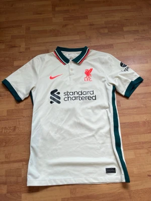 Liverpool bortatröja Nike 21/22 - Snygg Liverpool FC bortatröja från säsongen 21/22 i offwhite med gröna detaljer. Tröjan har tryck med Alexander-Arnold och nummer 66 på ryggen, samt klubbmärke och sponsorloggor framtill. Tillverkad i lätt och ventilerande material, perfekt för fotboll eller supporterliv.