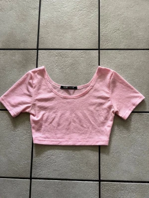 Rosa croppad topp från Now How storlek M/L - Söt ljusrosa croppad topp från Now How med rundad urringning och korta ärmar. Tillverkad i mjukt material som känns bekvämt mot huden. Perfekt att styla med höga jeans eller kjol för en trendig look.