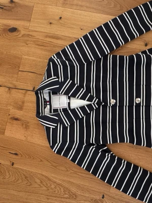 Randig kavaj från Tommy Hilfiger - Snygg kavaj från Tommy Hilfiger i marinblått med vita horisontella ränder. Klassisk krage, långa ärmar och silverfärgade knappar framtill. Perfekt för en trendig och stilren look. Materialet känns mjukt och bekvämt.