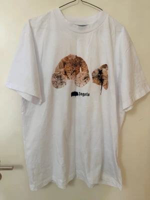 Vit t-shirt med björntryck - Säljer en vit t-shirt med ett unikt björntryck på bröstet och texten 'Angels' under. T-shirten har korta ärmar och en klassisk passform. Perfekt för dig som gillar streetwear och vill sticka ut med en cool design.