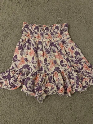 Blommig volangkjol i pastellfärger Pernilla Wahlgren  - Supersöt kortkjol med blommigt mönster i lila, rosa och vitt. Kjolen har bred smock i midjan och volang längst ner för extra flowy känsla. Perfekt för varma dagar och ger en romantisk vibe till din outfit.