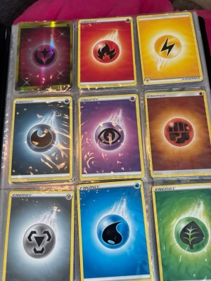 Pokémon energikort samling - Säljer en samling Pokémon energikort i olika färger och typer, bland annat eld, el, gräs, vatten, mörker, psykisk och metall. Kort med både vanliga och holo-effekter, perfekta för att bygga eller komplettera din lek. Alla korten är insatta i plastfickor. Fairy-type kortet med guldram.