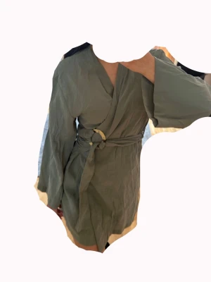 Olivgrön kimono klänning - Snygg olivgrön klänning i kimonomodell med knytband i midjan och vida ärmar. Har kontrastfärgade detaljer i beige vid ärmslut och nederkant. Perfekt klänning nu till sommar och superbekväm tack vare det mjuka materialet. Aldrig använd! OBS: FRAKTAR INOM 24H!🩷