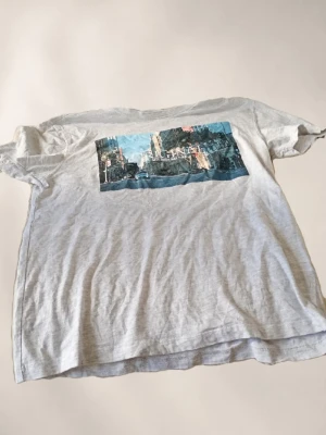 Ljusgrå t-shirt med tryck  - Ljusgrå t-shirt med ett rektangulärt tryck framtill som föreställer en stadsmiljö i blå och beige toner. Klassisk passform med korta ärmar och rund halsringning. Perfekt för en avslappnad och cool stil.
