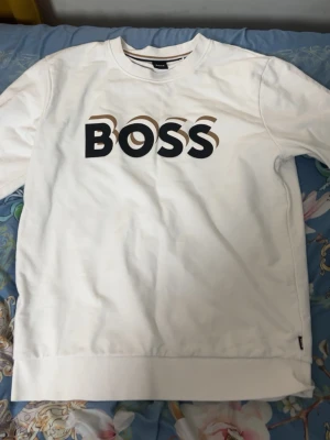 Vit sweatshirt från BOSS - Snygg vit sweatshirt från BOSS med stor logga i svart och beige framtill. Tröjan har rund halsringning och ribbade muddar vid ärmslut och nederkant. Perfekt för en clean och stilren look.