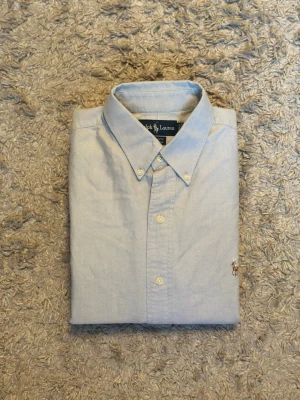 Ljusblå skjorta från Ralph Lauren - Klassisk ljusblå skjorta från Ralph Lauren med button-down krage och broderad logga på bröstet. Skjortan har långa ärmar, knappar framtill och är tillverkad i bomull. Perfekt för en clean och stilren look.