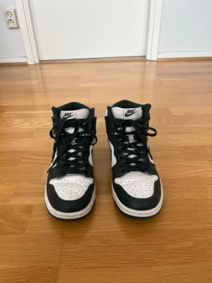 Nike Dunk High Sneakers - Storlek 41 - Säljer ett par Nike Dunk High i en stilren mörkgrön och vit färgkombination. Skorna är i bra använt skick med normalt slitage från användning, men har fortfarande mycket kvar att ge. En klassisk och lättmatchad sneaker som passar både till vardags och streetwear-stil. Storlek 41 (EU). Skorna är bekväma, välvårdade och redo för en ny ägare.