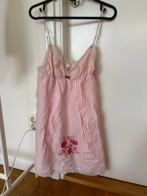 Rosa nattlinne från Odd Molly - Sött rosa nattlinne från Odd Molly med tunna axelband, spetsdetaljer och broderi i rött nertill. Liten rosett framtill och dekorativa veck på framsidan.