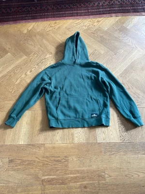 Grön hoodie med snowboard-tryck - Säljer en grön hoodie med ett litet snowboard-tryck på bröstet. Hoodien har en klassisk känguruficka framtill och ribbade muddar vid ärmar och nederkant. Perfekt för dig som gillar streetwear och sportig stil. Storlek 146-152