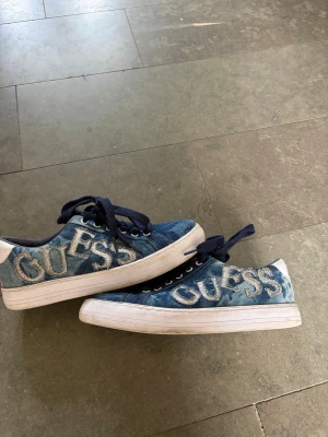 Blå denim Guess sneakers - Snygga sneakers från Guess i blått denimtyg med stora broderade logotyper på sidorna. Skorna har vita sulor och svarta skosnören. Perfekta för dig som gillar streetstyle och vill sticka ut med ett coolt märke.