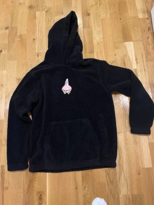 HM Svart fleece hoodie stl M  med Patrick från Svampbob - Mysig svart hoodie i teddy-material med huva och stor magficka. På bröstet finns ett broderat motiv av Patrick från Svampbob Fyrkant. Perfekt för dig som gillar tecknade serier och vill ha en skön och varm hoodie med unik detalj.
