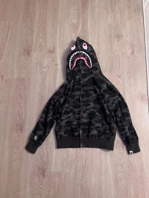 BAPE camo shark hoodie - Säljer en svart camo hoodie från BAPE med ikoniskt hajmotiv på huvan och broderade bokstäver 'WGM' i rött, blått och orange på sidan. Dragkedja framtill och ribbade muddar. Perfekt för dig som gillar streetwear och vill sticka ut.
