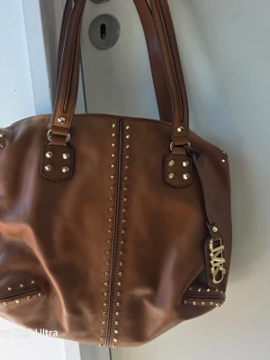 Brun handväska med nitar från Michael Kors - Snygg brun handväska i läder från Michael Kors med guldfärgade nitar och detaljer. Väskan har dubbla handtag och ett MK-hänge i metall. Perfekt för dig som gillar en trendig och stilren look med lite edge.
