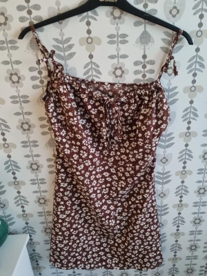 Brun blommig kortklänning med knyt - Söt brun kortklänning med vita blommor och knytband på axlarna. Klänningen har ett blommigt mönster och en avslappnad passform, perfekt för varma dagar. Materialet känns lätt och bekvämt.