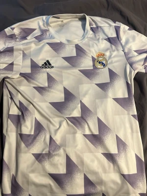 Real Madrid träningströja Adidas - Säljer en Real Madrid träningströja från Adidas i vitt med lila geometriskt mönster. Tröjan har klubbmärket på bröstet och Adidas-logga. Tillverkad i lätt och ventilerande material, perfekt för fotbollsträning eller gym.