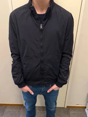 Woolrich windbreaker - Stilren svart vindjacka med hög krage och hel dragkedja framtill. Jackan har två sidofickor och muddar vid ärmslut och midja. Perfekt för blåsiga dagar och enkel att matcha med olika outfits.