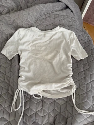 Vit ribbad topp med snörning - Säljer en vit ribbad topp med korta ärmar och snörning i sidorna. Toppen har en tight passform och är perfekt för att styla med jeans eller kjol. Materialet är mjukt och stretchigt, vilket gör den superbekväm.