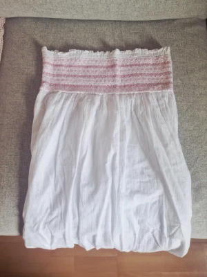 Vit sommarklänning från Gina Tricot - Söt vit klänning från Gina Tricot med smock upptill och rosa ränder. Klänningen är axelbandslös och har en luftig, lätt form som passar perfekt till varma dagar. Tillverkad i mjukt bomullstyg för en bekväm känsla.