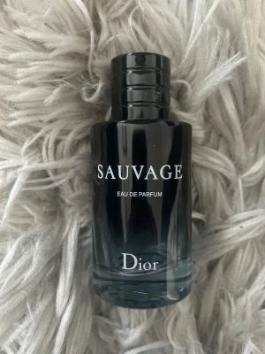 Dior Sauvage Eau de Parfum - Dior Sauvage Eau de Parfum i en elegant cylinderformad glasflaska med svart och transparent design. Flaskan har tydlig logga och text i vitt. En ikonisk doft som utstrålar lyx och modernitet, perfekt för dig som vill ha en klassisk men ändå fräsch parfym.